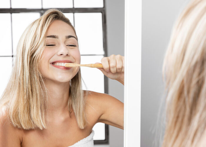 Young woman brushing teeth in front of a mirror, illustrating a low effort high reward daily habit for better health.