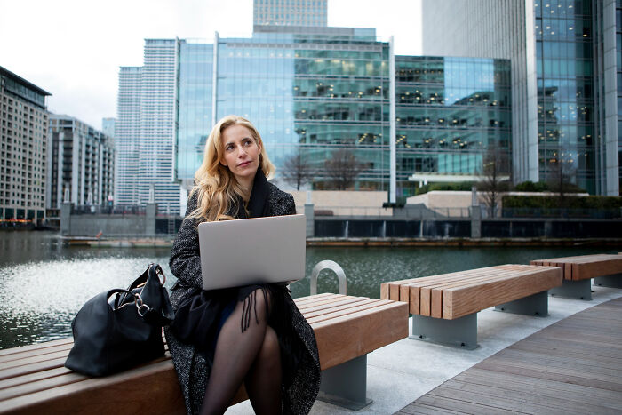 Woman working on laptop outdoors by city waterfront, reflecting on unhinged hacks done to land job successfully.