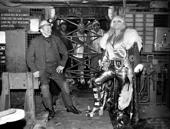Black and white historical photo showing miners and a person in shiny metallic outfit, highlighting unique historical moments.