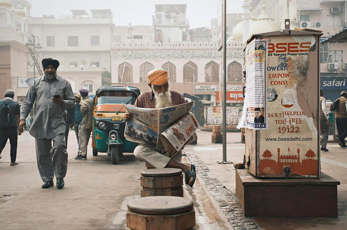 Man in a turban reading a newspaper on a busy street in Asia, capturing candid everyday moments by photographer Gil Kreslavsky