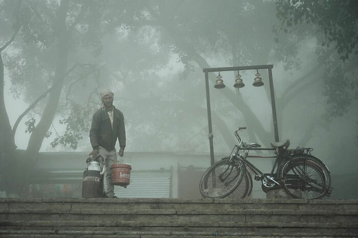 Man carrying milk cans on foggy street beside bicycle and bells, candid everyday moment from Asia captured by photographer.