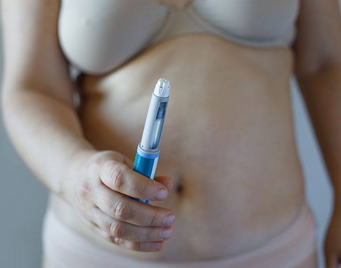 Person holding a blue and white injection pen in front of bare midsection, related to Ozempic vulva side effects. Person holding a blue and white injection pen in front of bare midsection, related to Ozempic vulva side effects.
