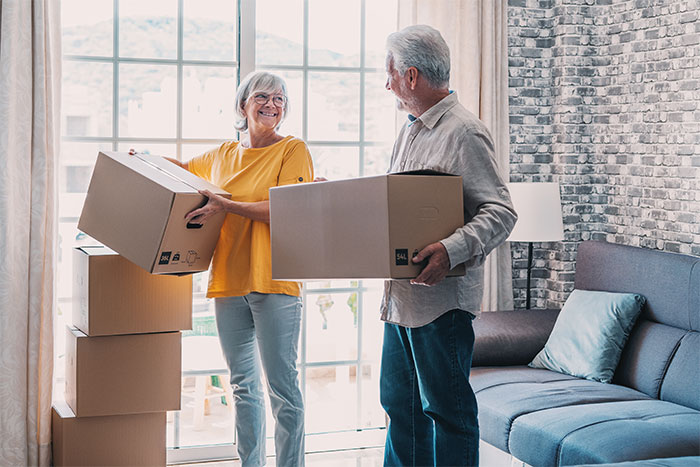 Elderly couple smiling and carrying moving boxes inside a living room, representing parents visiting their son's home. Elderly couple smiling and carrying moving boxes inside a living room, representing parents visiting their son's home.