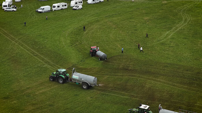 Aerial view of a farmer spraying manure with a tractor near squatters’ caravans on a large grassy field. Aerial view of a farmer spraying manure with a tractor near squatters’ caravans on a large grassy field.