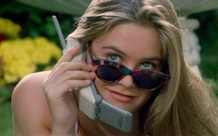 Alicia Silverstone wearing sunglasses and smiling while holding a large vintage cellphone outdoors on a sunny day