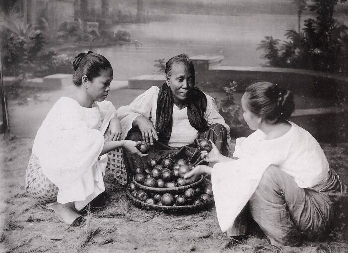 Three Filipino women in traditional attire sitting on the ground with baskets of fruit in a 1890s Philippines scene.