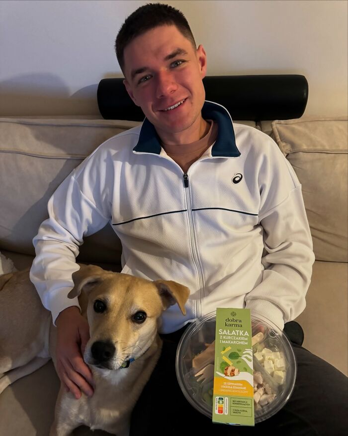 Young man wearing white jacket sitting with dog, holding salad pack, related to Polish CEO US Open hat snatcher news. Young man wearing white jacket sitting with dog, holding salad pack, related to Polish CEO US Open hat snatcher news.