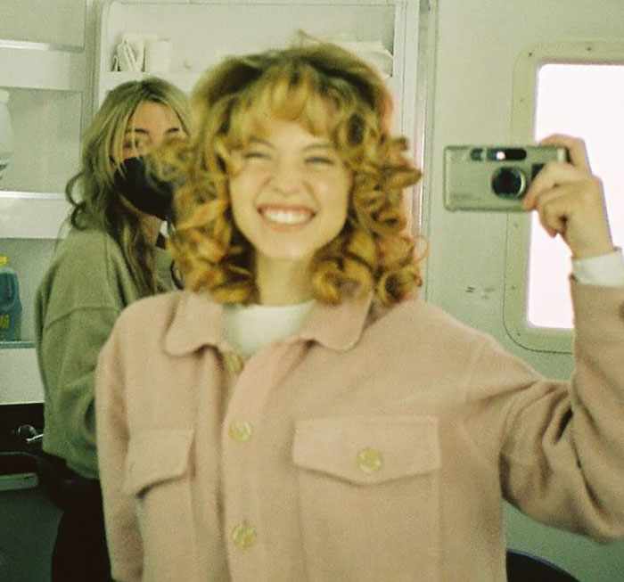 Sydney Sweeney smiling and taking a selfie indoors while another person wearing a mask stands in the background. Sydney Sweeney smiling and taking a selfie indoors while another person wearing a mask stands in the background.