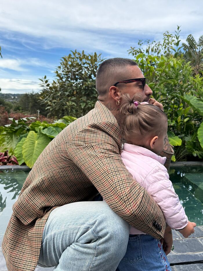 Man with sunglasses and tattoo hugging a toddler girl outdoors near plants, reflecting singer stopping concert over mom behavior. Man with sunglasses and tattoo hugging a toddler girl outdoors near plants, reflecting singer stopping concert over mom behavior.