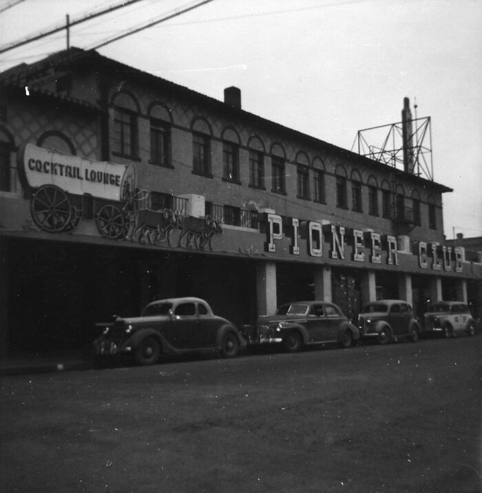 Historic black and white photo of early Vegas street with vintage cars parked outside Pioneer Club and cocktail lounge.