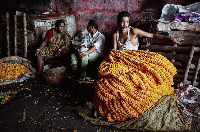 Three people in a rustic setting with large piles of orange marigold garlands, capturing candid everyday moments from Asia.