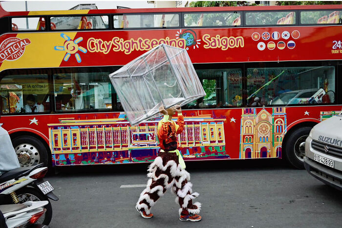 Person carrying a large transparent box on their head in a busy Asian street with colorful sightseeing bus in background
