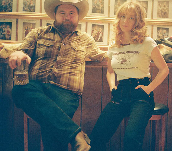 Man in cowboy hat with drink next to Sydney Sweeney wearing casual outfit in a vintage-style room setting. Man in cowboy hat with drink next to Sydney Sweeney wearing casual outfit in a vintage-style room setting.