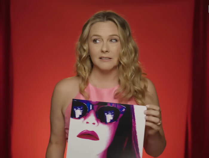 Woman in pink dress holding a stylized photo of Alicia Silverstone wearing sunglasses against a red backdrop