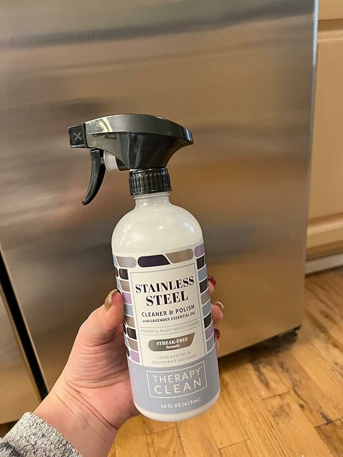 Hand holding a stainless steel cleaner spray bottle in front of a clean stainless steel appliance for genius cleaning tricks.