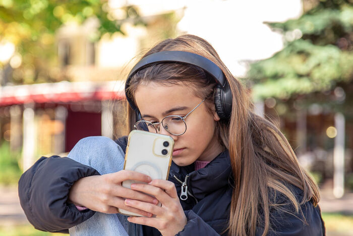 Teen girl wearing headphones and glasses focused on her phone, representing shopping and non-substance dependencies outdoors.