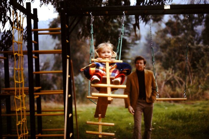 Child in colorful jacket enjoying a swing while an adult watches nearby, capturing candid glimpses into the past.