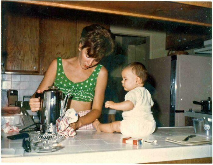 Woman in a green top pouring coffee while a baby sits on the kitchen counter in a candid glimpse into the past.