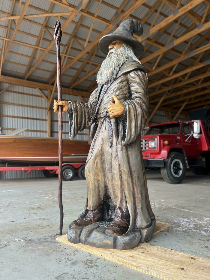 Woodworker’s detailed carving of a wizard holding a staff inside a large workshop with a red truck in the background.