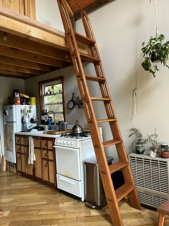 Woodworkers crafted a custom wooden ladder leading to a loft in a cozy kitchen with natural wood accents and plants.