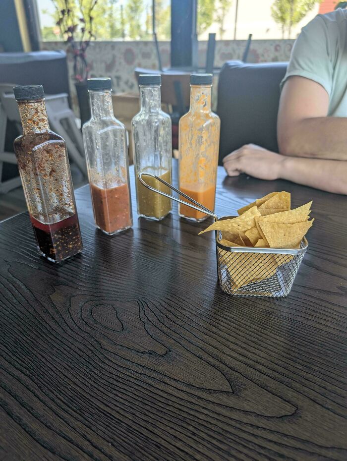 Small metal basket with tortilla chips served alongside four clear sauce bottles on a restaurant table, showcasing ridiculous food serving.