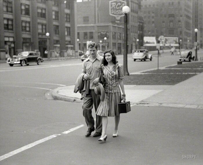 Couple walking on city street in vintage attire, capturing candid glimpses into the past with classic cars in background.