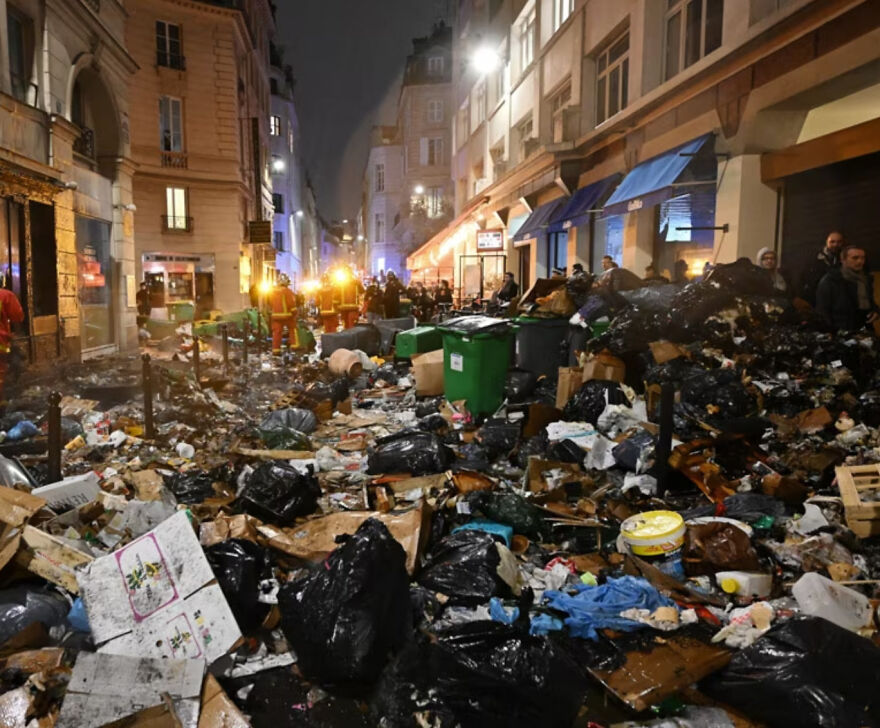 Street covered in piles of garbage bags and trash with people and lit buildings in the background at night in an urban area