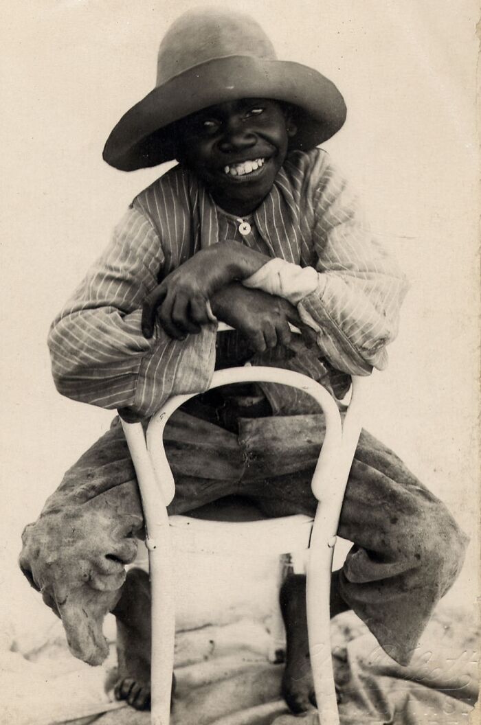 Young Indigenous Australian boy in early 1900s attire sitting on a chair, smiling in a rare historical photo glimpse.