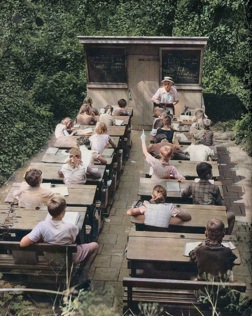 Outdoor classroom with children sitting at wooden desks, a teacher instructing, showcasing interesting historical school setup.