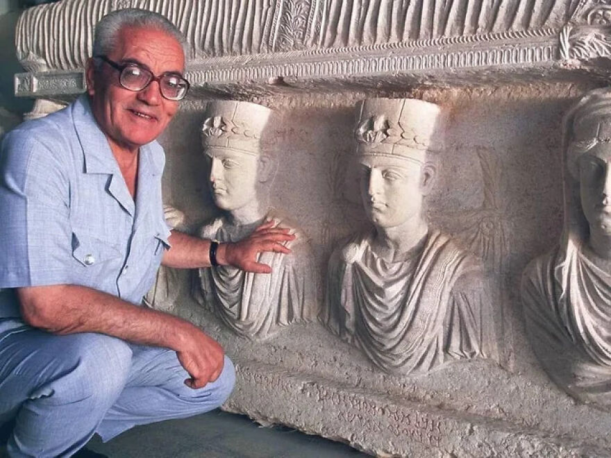 Man in glasses and light blue outfit examining ancient stone busts carved into a wall in an interesting historical setting.