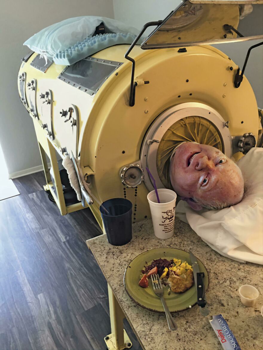 An elderly man inside a vintage medical oxygen chamber beside a table with food and drinks in an interesting online community post.