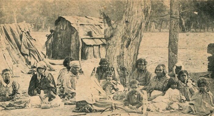 Group of Indigenous Australians from the 1900s sitting outdoors near traditional shelters and trees in a historical photo.
