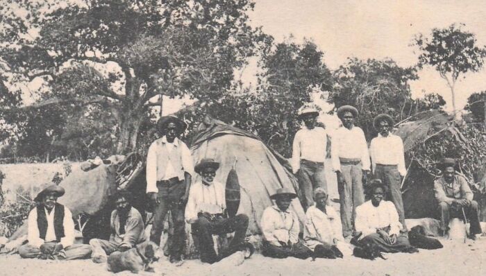 Group of Indigenous Australians from the 1900s dressed in traditional and period clothing, posing outdoors near shelters and trees.