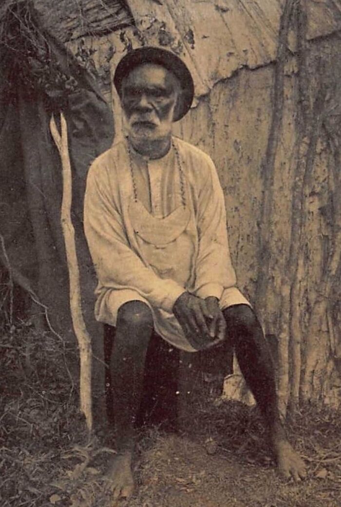Indigenous Australian elder from the 1900s sitting barefoot outdoors wearing traditional attire and necklace.
