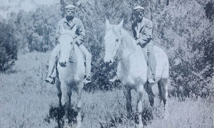Two Indigenous Australian men from the 1900s riding white horses in a natural, grassy landscape with sparse trees.