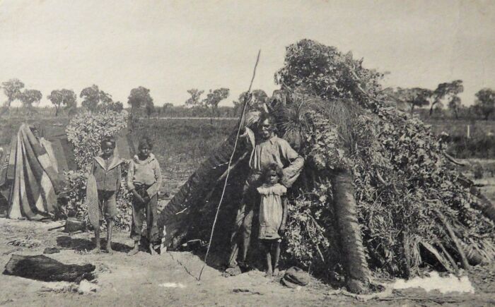 Indigenous Australians family standing outside traditional shelter in early 1900s, rare glimpses into historic daily life.