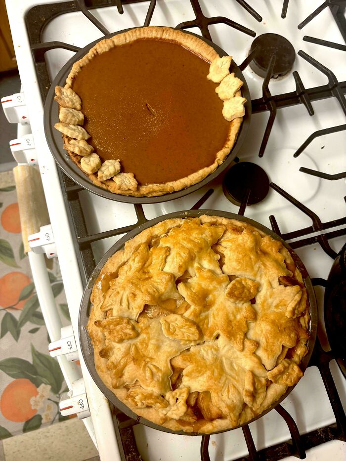 Two homemade baked goods on a stove, including a decorated pumpkin pie and a leaf-topped fruit pie.