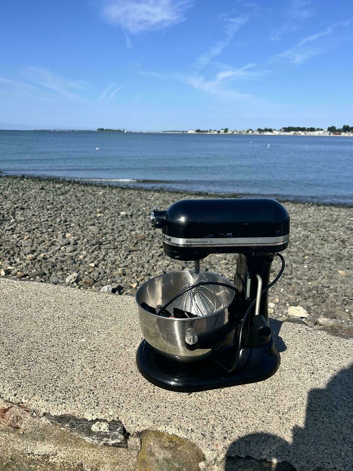 Black vintage stand mixer thrift store find placed on a concrete ledge with rocky beach and sea in the background.