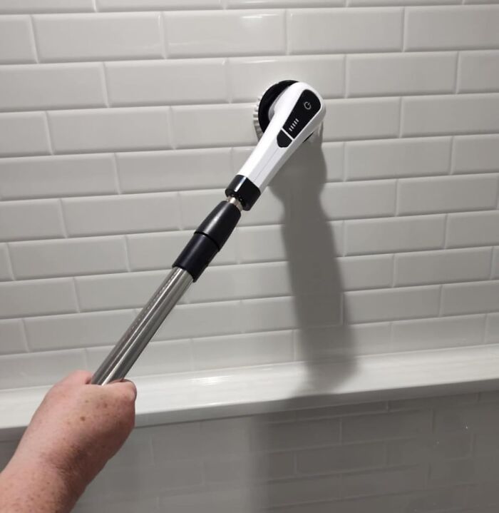 Hand holding a cleaning brush with an extendable handle scrubbing white tiled wall using genius cleaning tricks.