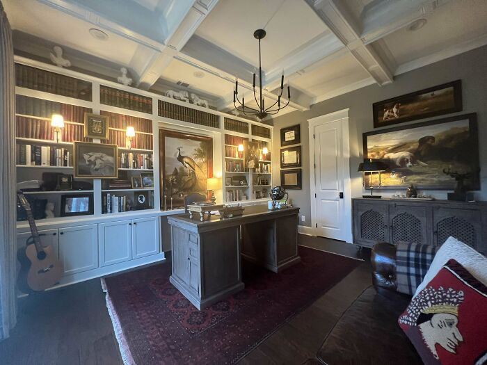 Cozy home office corner with wooden desk, built-in bookshelves, decorative lighting, and classic artwork on walls.
