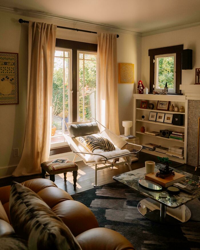 Cozy corner of home with natural light, decorated with patterned pillows, a chair, and bookshelf featuring art and books.