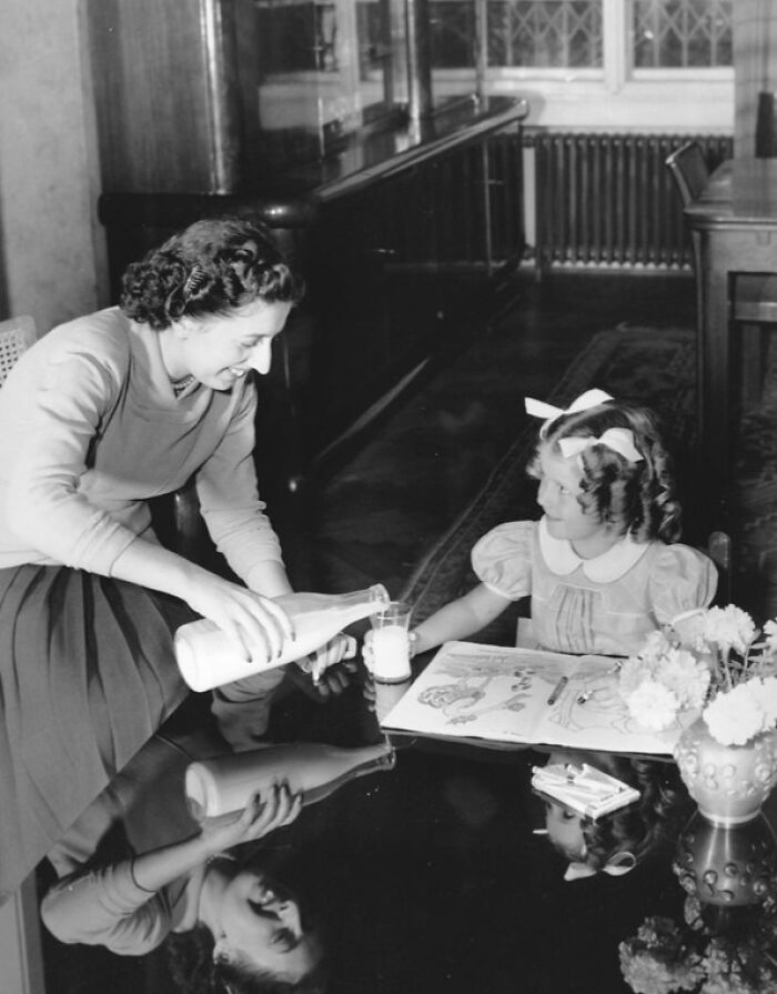 Woman pouring milk into a glass for a young girl sitting at a table with a coloring book rare WWII photos.