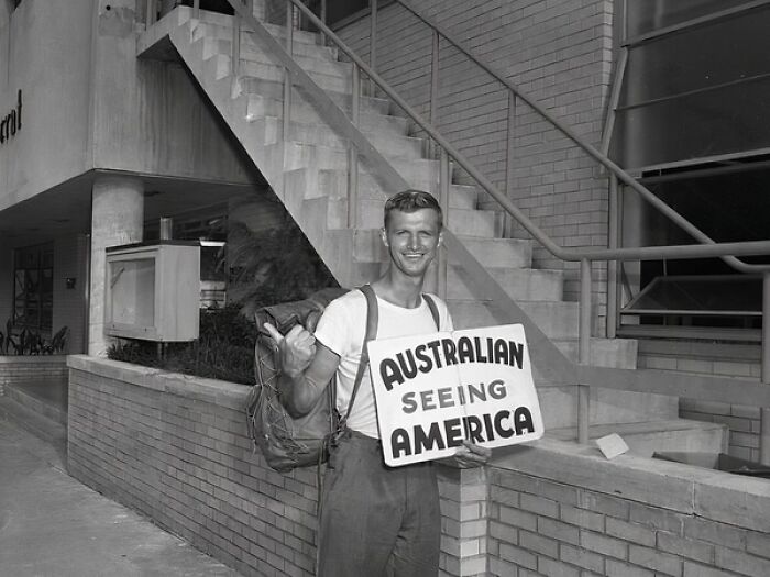 Young man hitchhiking with a backpack holding a sign in a street scene from everyday life in 1950s Florida.