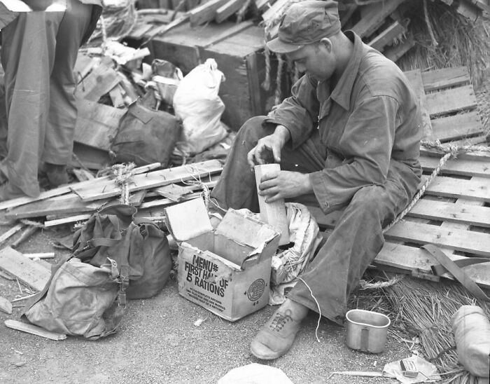 WWII soldier sitting among supplies, opening a box of first aid and rations in a rare WWII photo from the era.