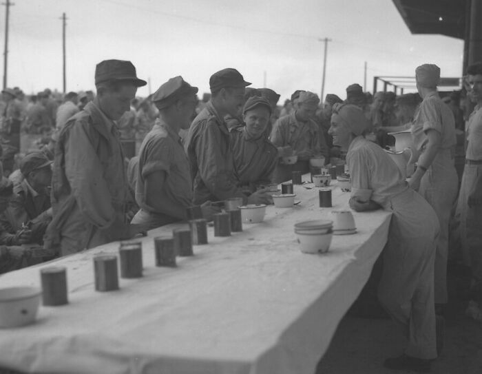 Rare WWII photo of soldiers in uniform lining up for meals served by a female worker at an outdoor military canteen.