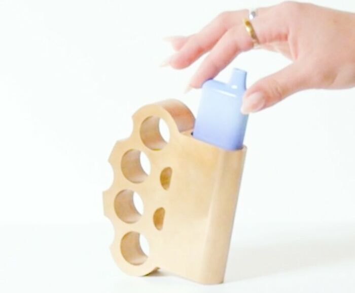 Hand holding a blue vape device near a wooden accessory shaped like brass knuckles, showcasing unusual accessories by Nik Bentel.