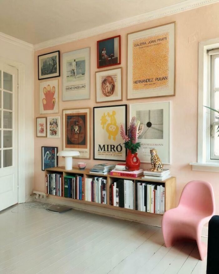 Cozy corner of a home with framed art, floating bookshelf filled with books, red vase with flowers, and a modern pink chair.