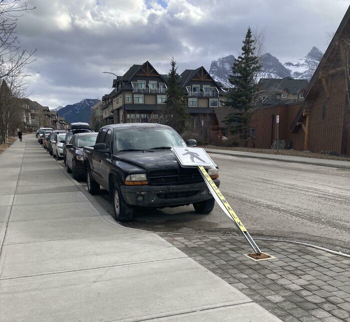 Black pickup truck parked with a fallen pedestrian sign leaning against it on a cloudy day in a mountain town.