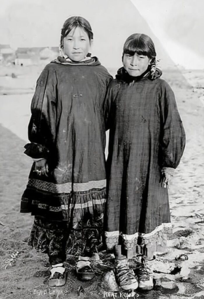 Two young Inuit girls standing side by side in traditional clothing in a historic black and white photo.