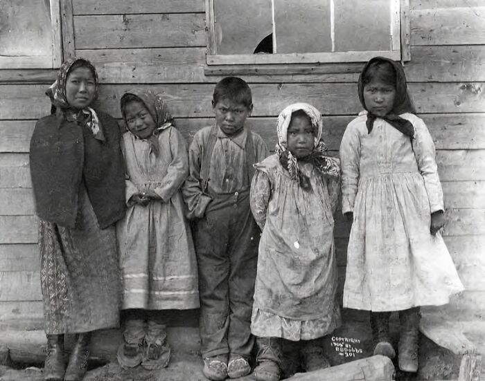 Historic black and white photo of Inuit children standing in front of a wooden building, capturing Inuit spirit and resilience.
